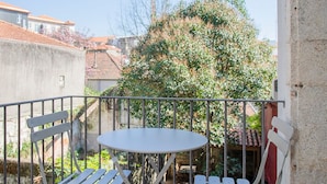 Ein Balkon mit rundem Tisch und zwei Klappstühlen, einem Metallgeländer sowie Blick auf einen Baum und Gebäude im Hintergrund.