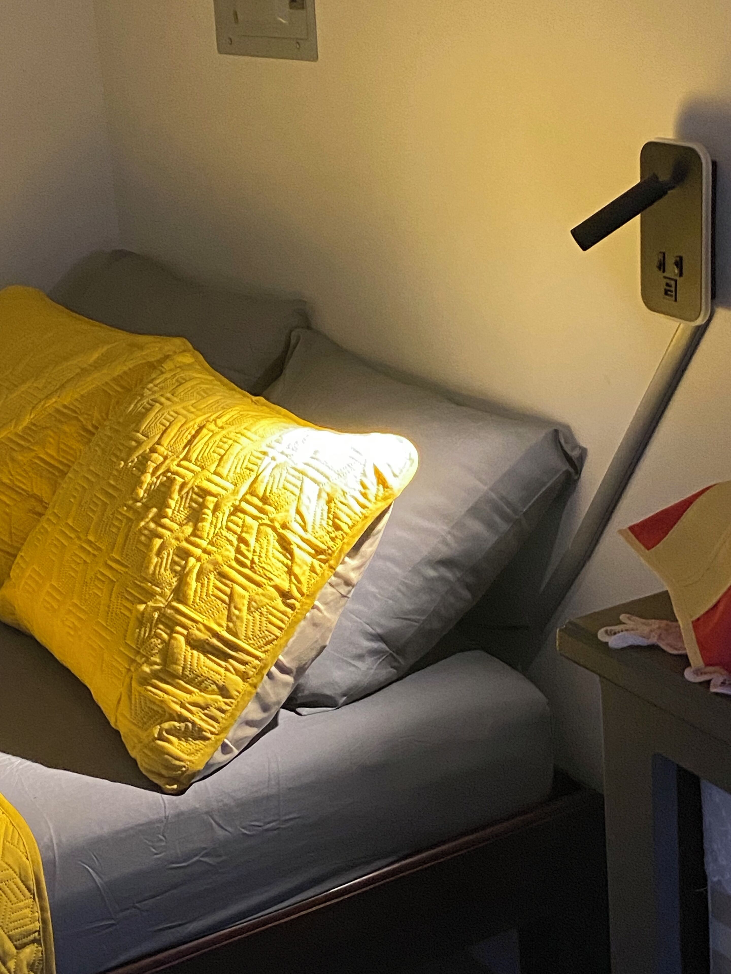 Un lit avec un oreiller jaune et un oreiller gris, une table de chevet avec une lampe et une prise électrique au mur.