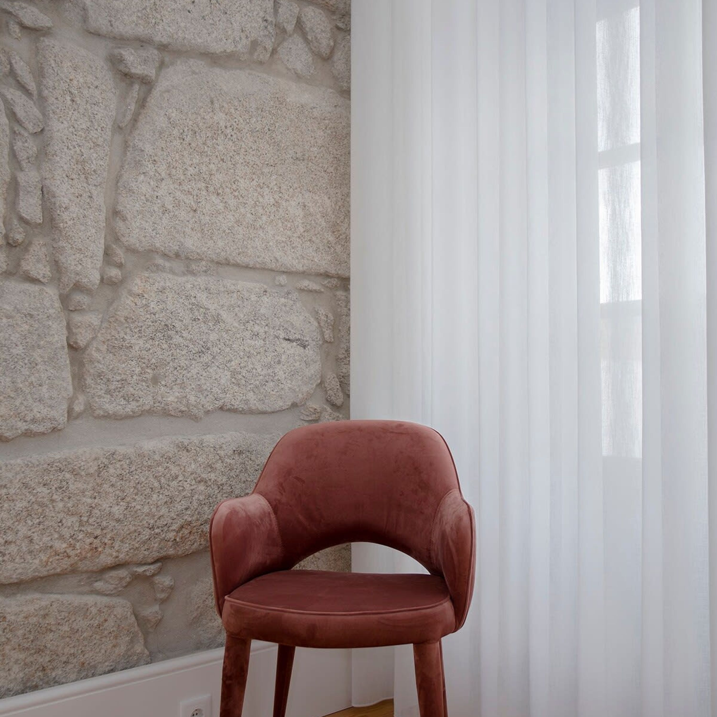 A room with a stone wall, a window with sheer curtains, and a single brown chair.