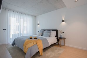 A modern bedroom with a large bed, a bedside table with a vase, and a window with sheer curtains. - Liiiving in Gaia - Beach Seaside Apartment (Vila Nova de Gaia)