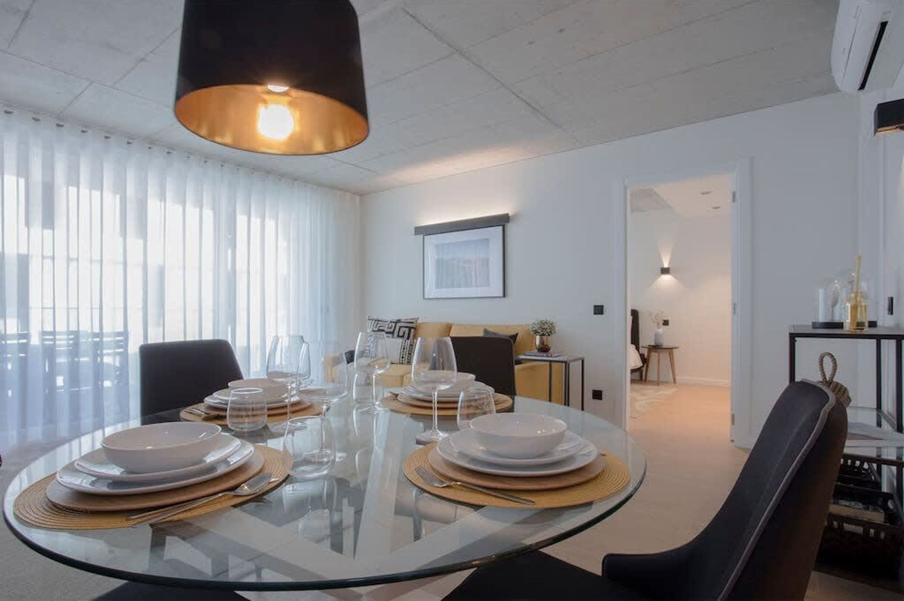 Un espace repas moderne avec une table ronde en verre, dressée pour quatre personnes, des assiettes blanches et de la verrerie transparente.