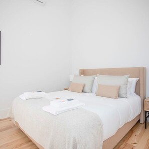 A neatly made bed with white linens and beige pillows, a wooden nightstand with a lamp, and a white wall. - Liiiving in Porto City View Apartment 1F (Porto)