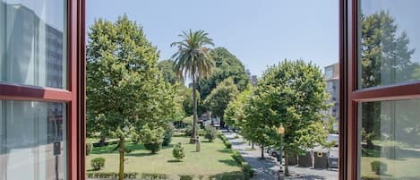Vista desde un balcón con barandilla de hierro forjado, que da a un jardín bien cuidado con árboles y una palmera, y una calle al fondo.