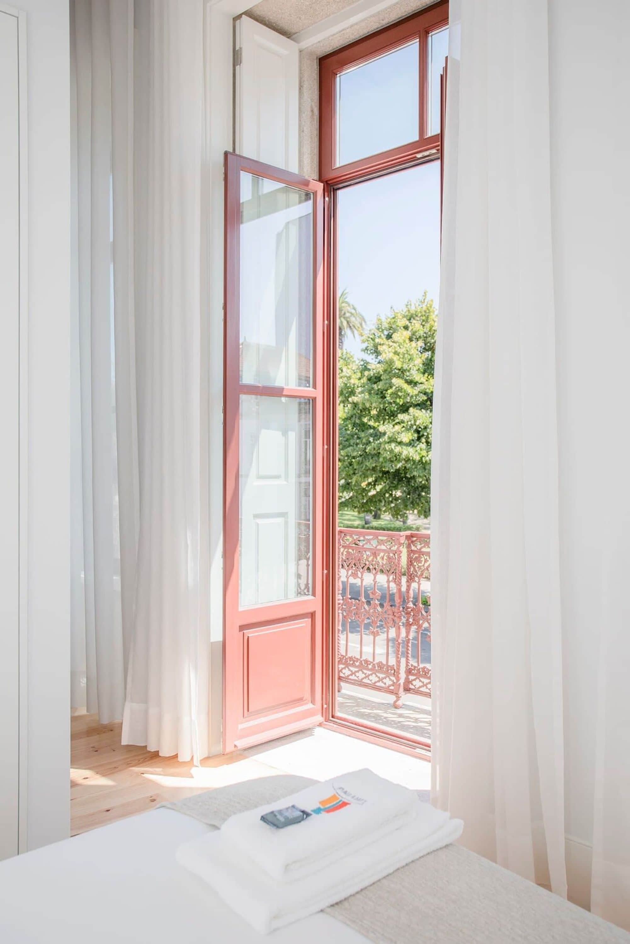 A room with a large window, white curtains, a wooden floor, and a neatly made bed with a white blanket and a folded towel.