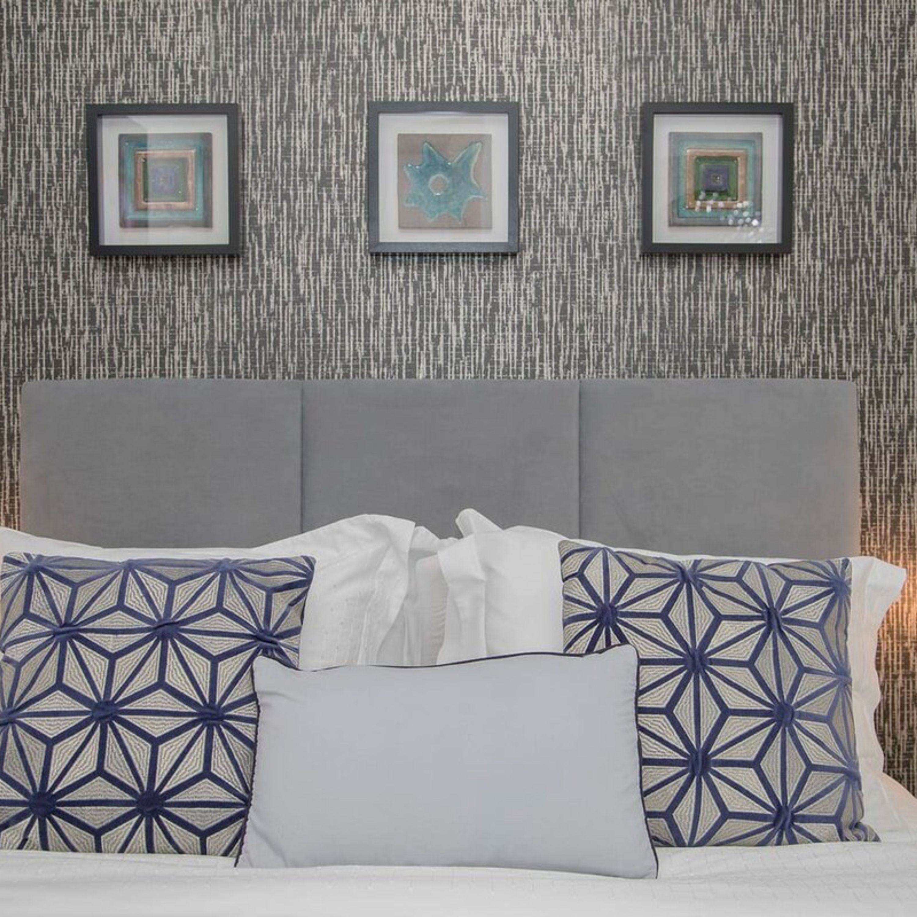 A bed with patterned pillows, a grey headboard, and three framed artworks on the wall.