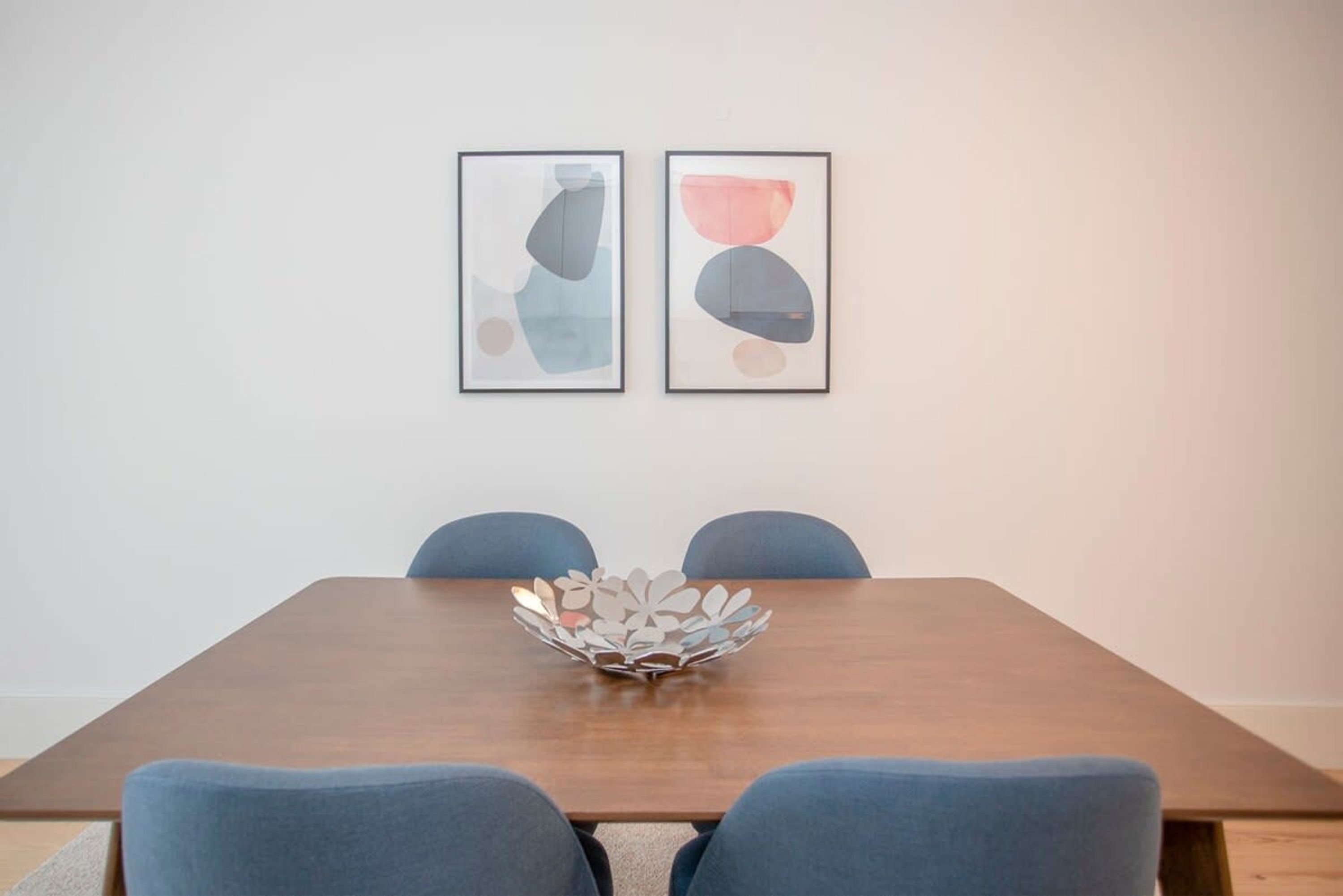A dining table with four chairs, a decorative metal bowl, and two abstract paintings on the wall.