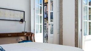 Ein Schlafzimmer mit Bett, Schreibtisch, Stuhl, Lampe und einem großen Fenster mit Blick auf ein Gebäude.