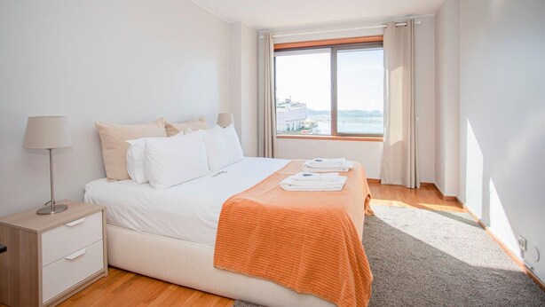 A hotel room with a large bed, a nightstand, a lamp, and a window with a view of the sea. - Liiiving in Porto Sea River View Apartment (Porto)