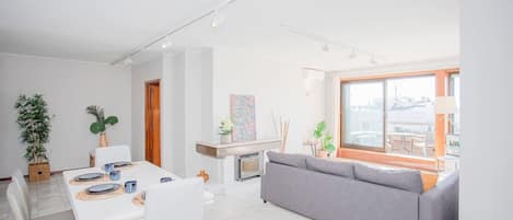 A modern living room with a grey sofa, a dining table with white chairs, and a kitchen area in the background.