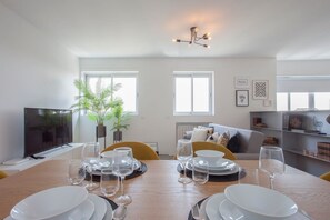 Un coin repas avec une table en bois dressée pour le repas, un téléviseur sur la gauche et un canapé au fond.
