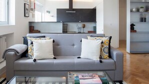 Ein modernes Wohnzimmer mit einem grauen Sofa, einem Glastisch und einer Küche im Hintergrund.