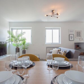 Un coin repas avec une table dressée pour quatre personnes, deux fenêtres, une plante et un canapé à l’arrière-plan.