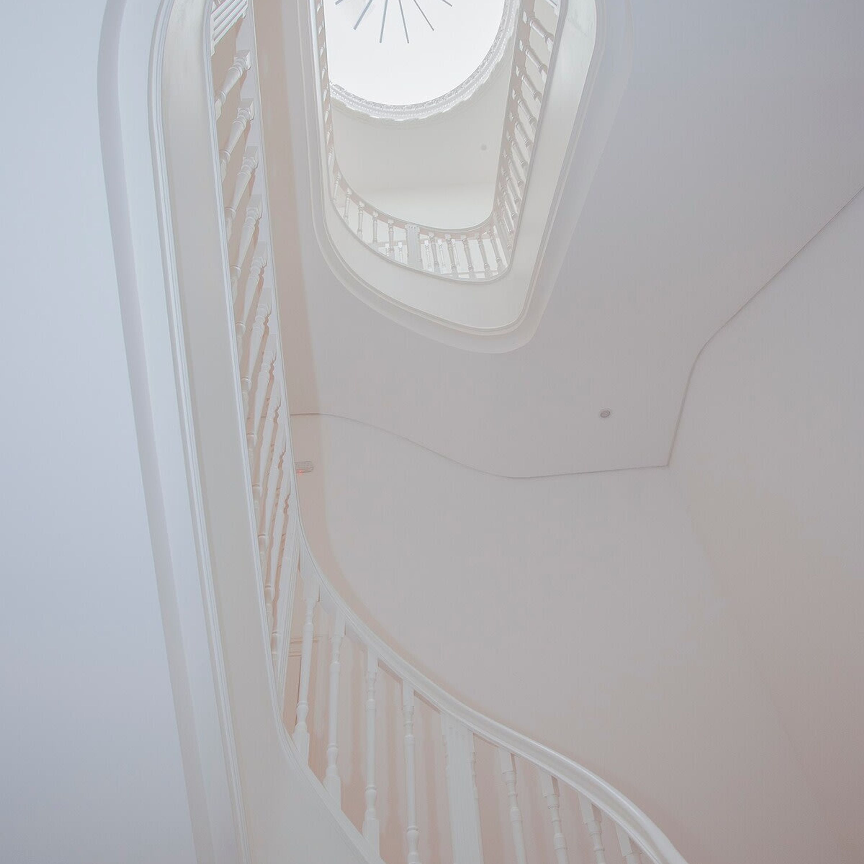 Una escalera en espiral con un espacio abierto en el centro, paredes blancas y un tragaluz circular en el techo.