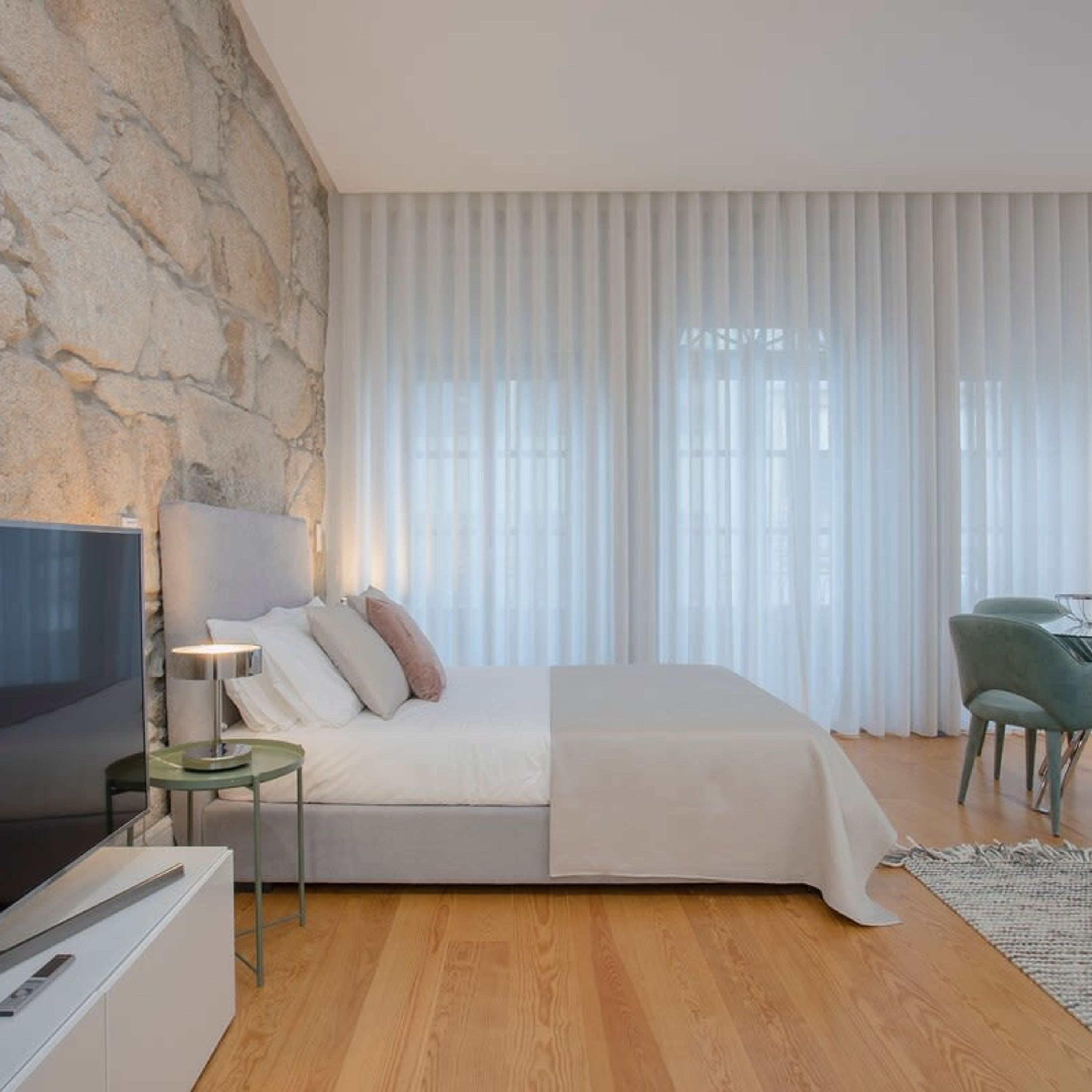 A modern bedroom with a stone wall, a bed with white bedding, a TV on a white cabinet, and a desk with a chair.