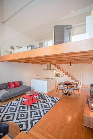 Um apartamento tipo loft moderno, com pé-direito alto, escada que leva a um andar superior, cozinha e área de estar com sofá e mesa de jantar.