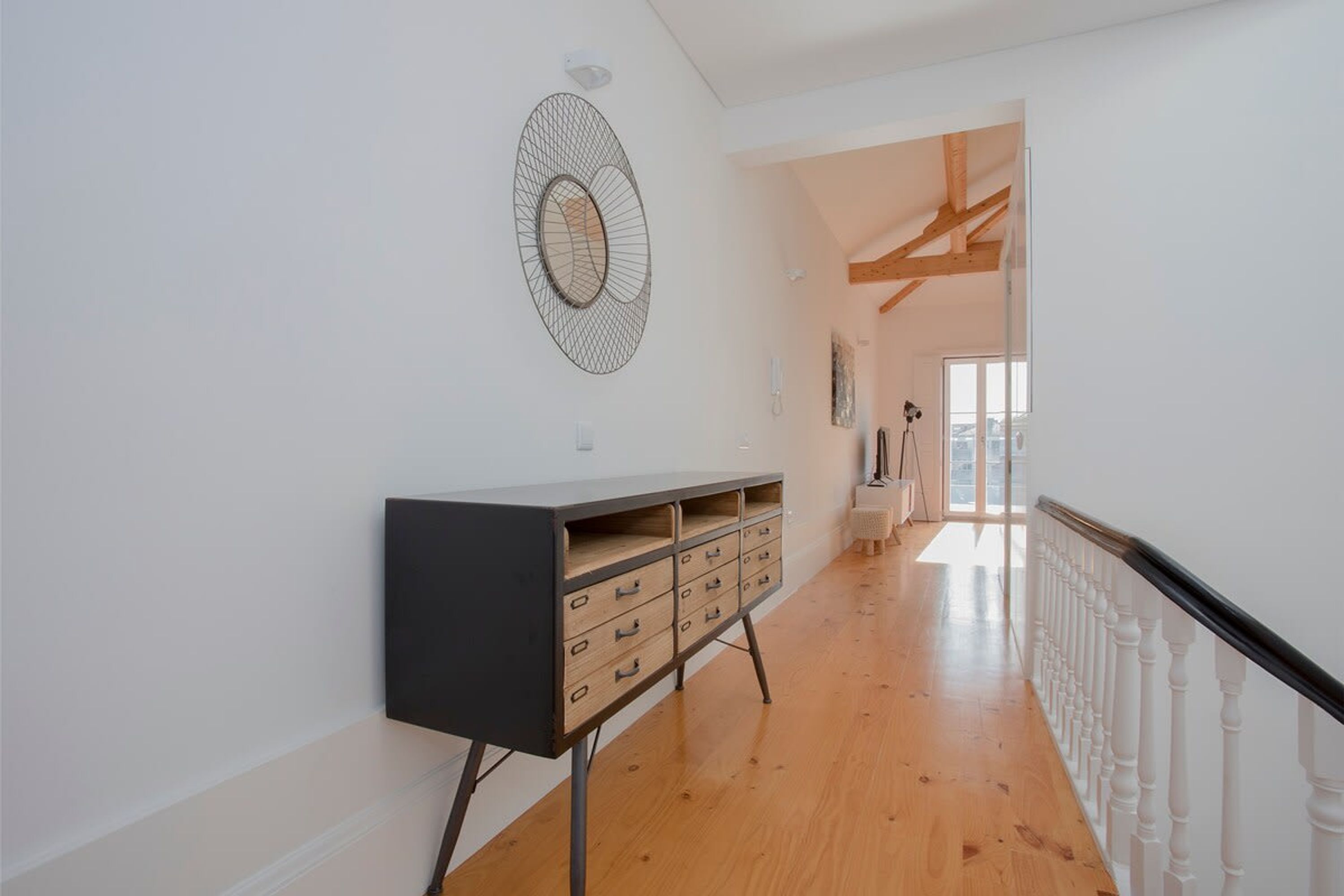 A hallway with a wooden floor, a black and wooden cabinet, white walls, and a white railing.