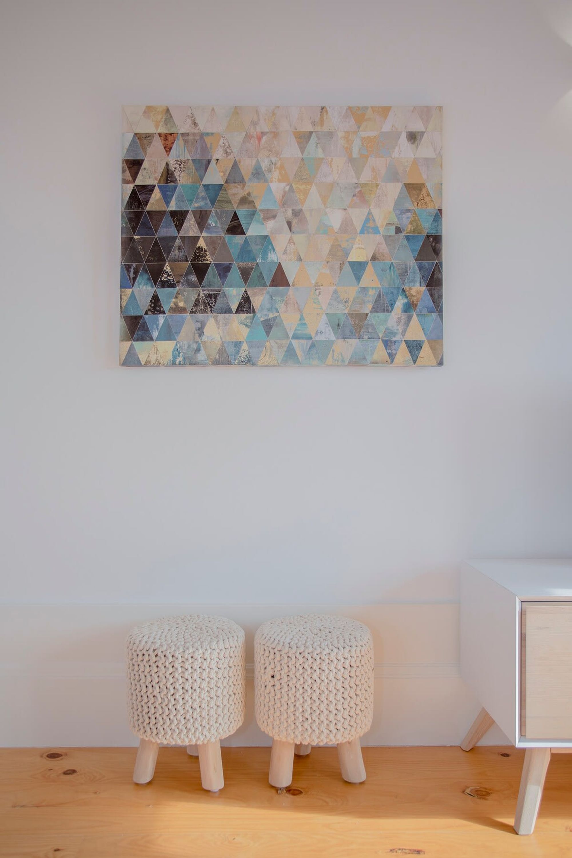 A room with a white wall, a geometric patterned artwork, and two white knitted stools.