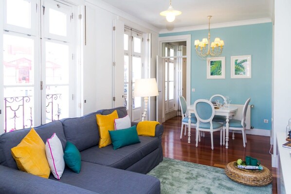 A living room with a grey sofa, teal and yellow cushions, a dining table with chairs, a chandelier, and a view through large windows. - Liiiving in Porto Ribeira Boutique Apartment (Porto)