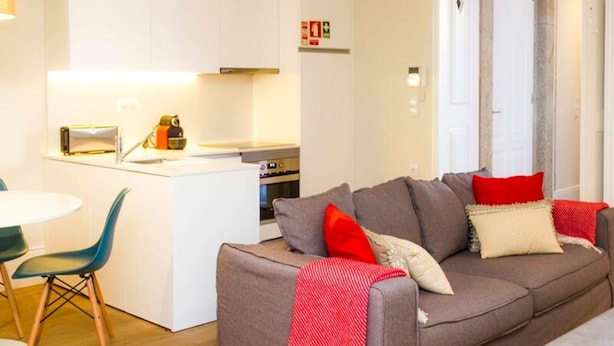 A modern living room with a grey sofa, red and white cushions, a yellow rug, and a small dining table with a green chair. - Liiiving in Porto Central Garden Flat (Porto)