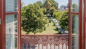 Une vue depuis un balcon orné d’une balustrade en fer forgé, donnant sur un jardin bien entretenu avec des arbres et un palmier, et sur une vue de la ville en arrière-plan.