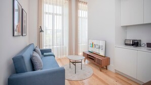 Un salon moderne avec un canapé bleu, une petite table ronde, un téléviseur et un coin cuisine équipé de placards et d’appareils électroménagers.