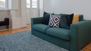 Ein Wohnzimmer mit einem türkisfarbenen Sofa, einem gemusterten Zierkissen und einem blauen Teppich mit Struktur.