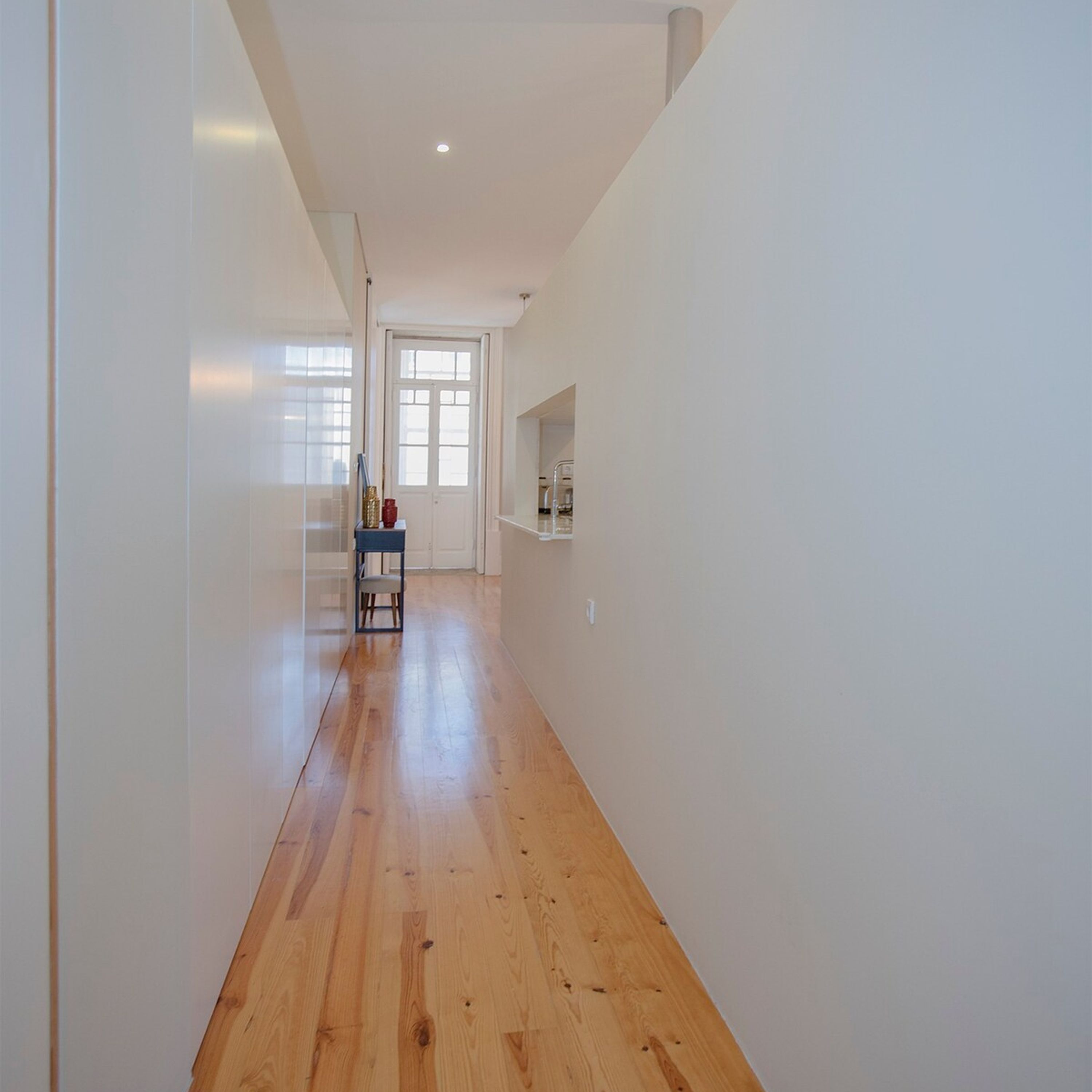 A hallway with wooden flooring, white walls, and a door leading to another room.