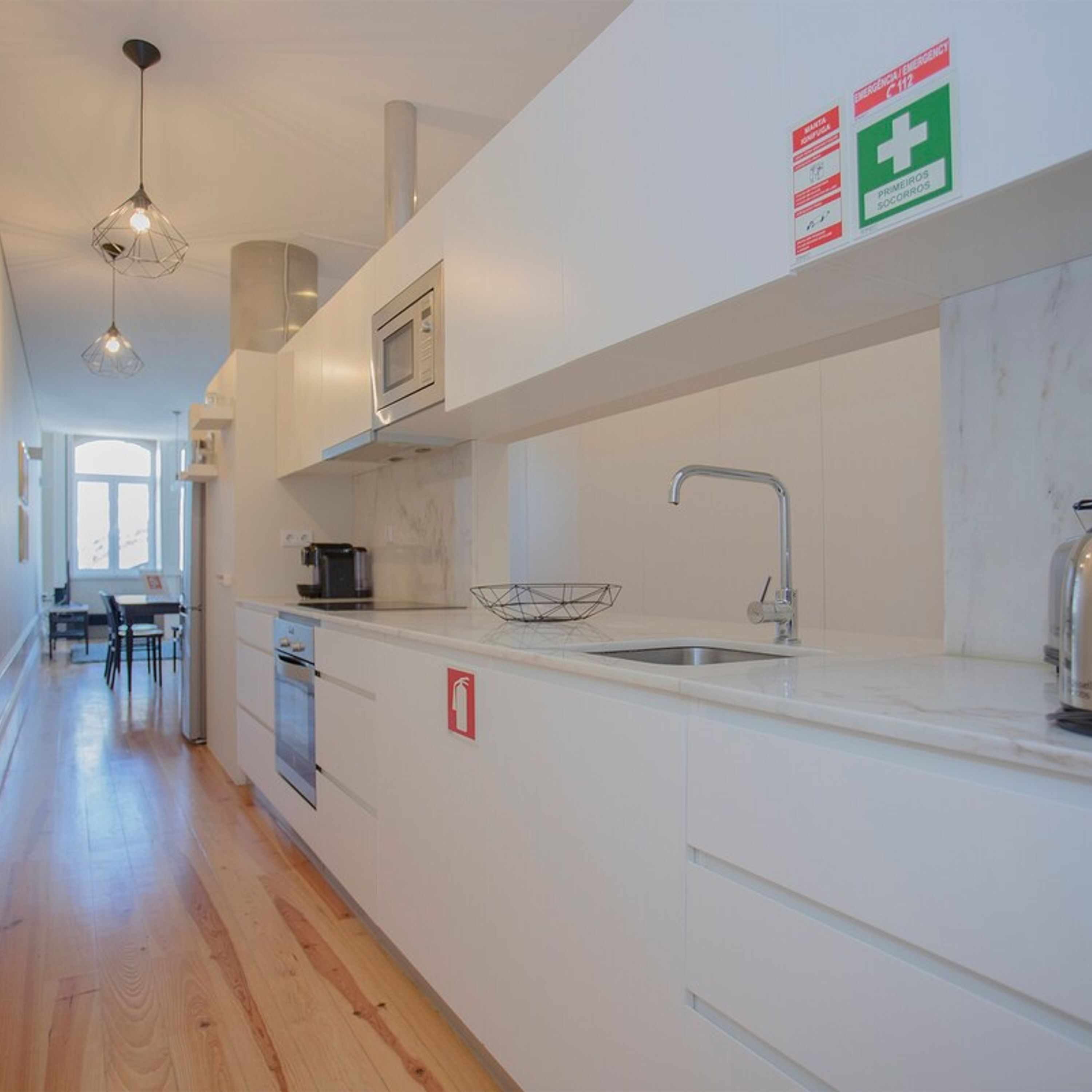 A modern kitchen with white cabinetry, a stainless steel sink, and a microwave. There are emergency signs on the wall and a fire extinguisher on the counter.