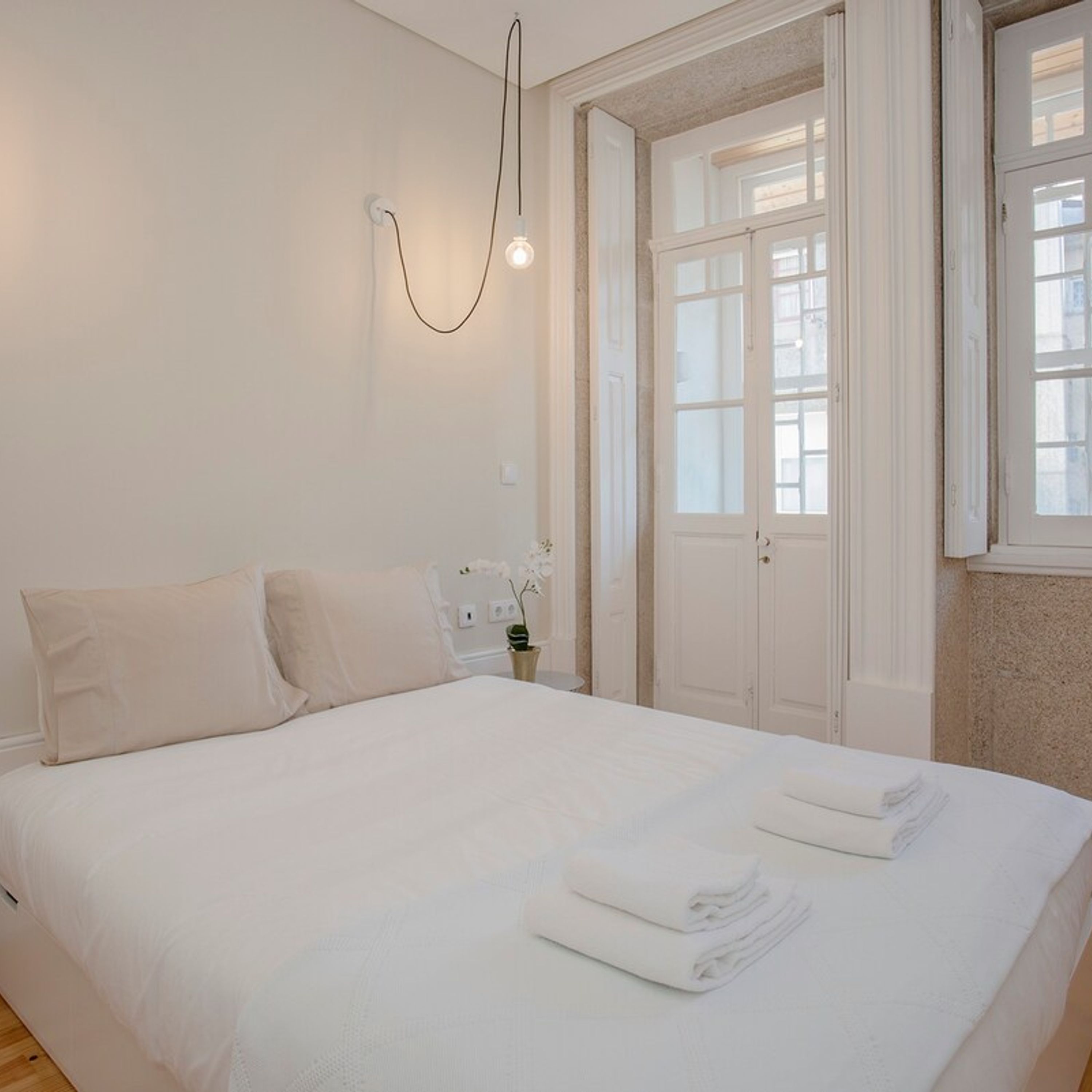 Un lit bien fait, avec du linge de lit blanc, une petite table de chevet ornée d’une plante et une porte dont les panneaux sont en verre dépoli.