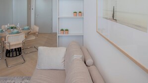 Ett modernt vardagsrum med en beige soffa, en matplats med ett bord för fyra personer och en bokhylla med små växter.