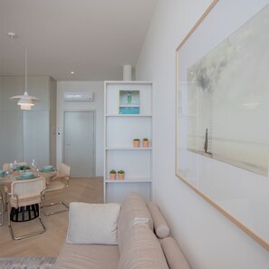 A modern living room with a beige sofa, a dining area with a table set for four, and a bookshelf with small plants. - Liiiving in Porto Luxury Beachfront Apartment II (Porto)