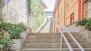 A set of stone steps with a metal handrail, flanked by buildings with a stone wall covered in ivy and a red building with a window. - Liiiving in Porto - Ribeira Riverside Apartment (Porto)