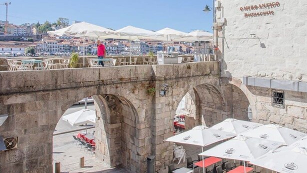 A stone bridge with arches, a white building with a restaurant sign, and outdoor seating with umbrellas. - Liiiving in Porto - Ribeira Riverside Apartment (Porto)