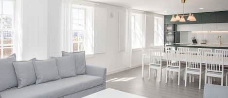 Uma sala moderna com um sofá cinzento, uma mesa de centro branca com um tabuleiro dourado, e uma área de cozinha com armários brancos e um conjunto de jantar.