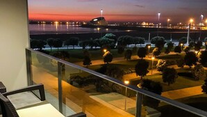 A balcony with a view of a cityscape at dusk, featuring a modern building, street lights, and a well-maintained garden. - Liiiving in Matosinhos - High Sea Condo (Matosinhos)