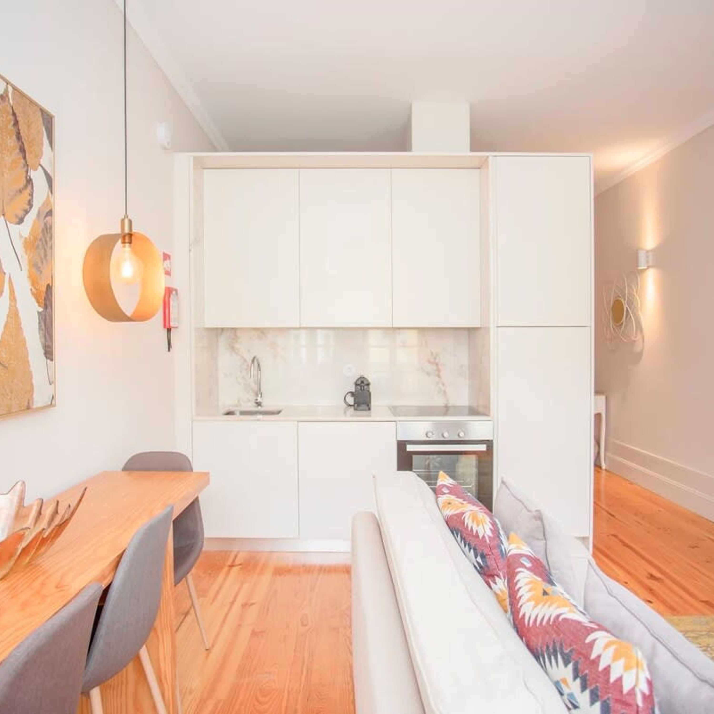 Eine kompakte Küche mit weißen Schränken, einem Esstisch mit Stühlen und einem Sofa mit farbigem Kissen.