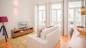 A modern living room with a sofa, a TV on a wooden console, and large windows with a view of buildings. - Liiiving in Porto - Ribeira Secret Apartment (Porto)