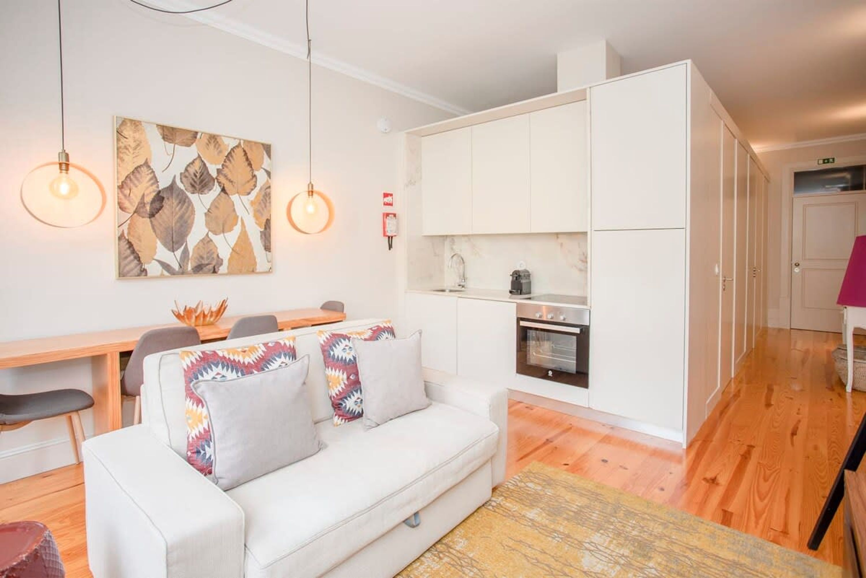 A modern living room with a white sofa, a dining table, and a kitchen area with white cabinets and a stove.