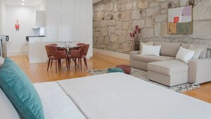 A modern living room with a stone wall, a sofa, a dining area with chairs, and a kitchen in the background. - Liiiving in Porto Downtown Luxury Apartment 2T (Porto)