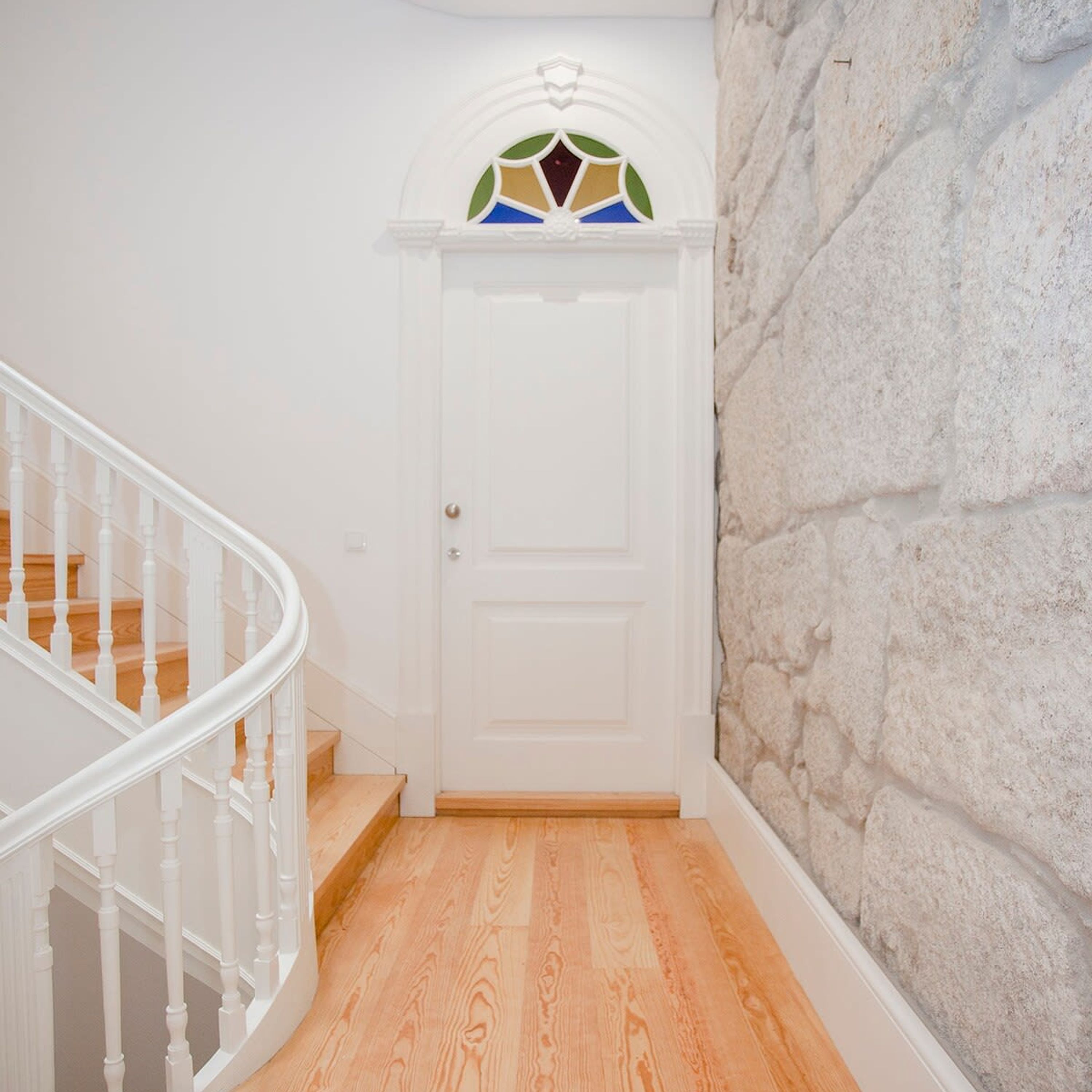 A white door with a stained glass window above it, a wooden staircase with white balusters, and a textured stone wall.