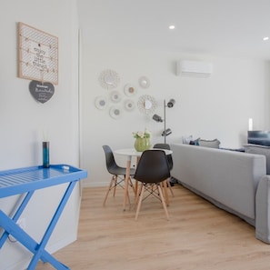 A modern living room with a grey sofa, a small dining table with chairs, a blue folding table, decorative wall art, and a ceiling-mounted air conditioner. - Liiiving in Porto Bright Light Studio (Porto)