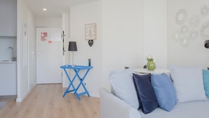 A modern living room with a grey sofa, a blue coffee table, and a small white table with a vase. There is a kitchen area in the background with a white door and a blue folding table. - Liiiving in Porto Bright Light Studio (Porto)