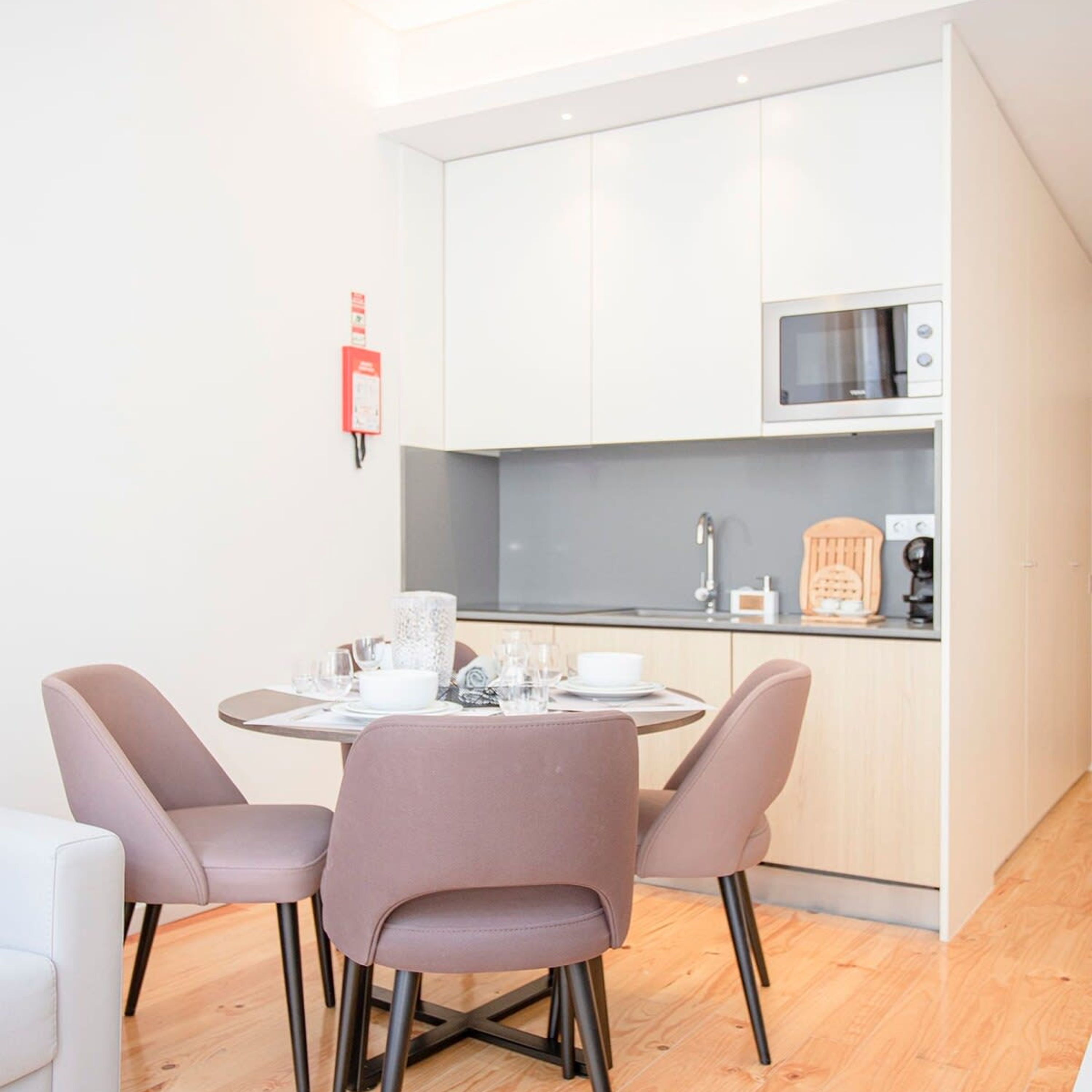 A compact kitchen with white cabinets, a microwave, and a sink. There is a dining table with chairs and a fire extinguisher mounted on the wall.