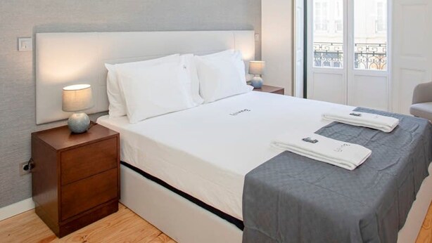 A modern hotel room with a large bed, a nightstand, a lamp, and a view of the city through the window. - Liiiving in Porto-mouzinho Residence Apartment 1F (Porto)
