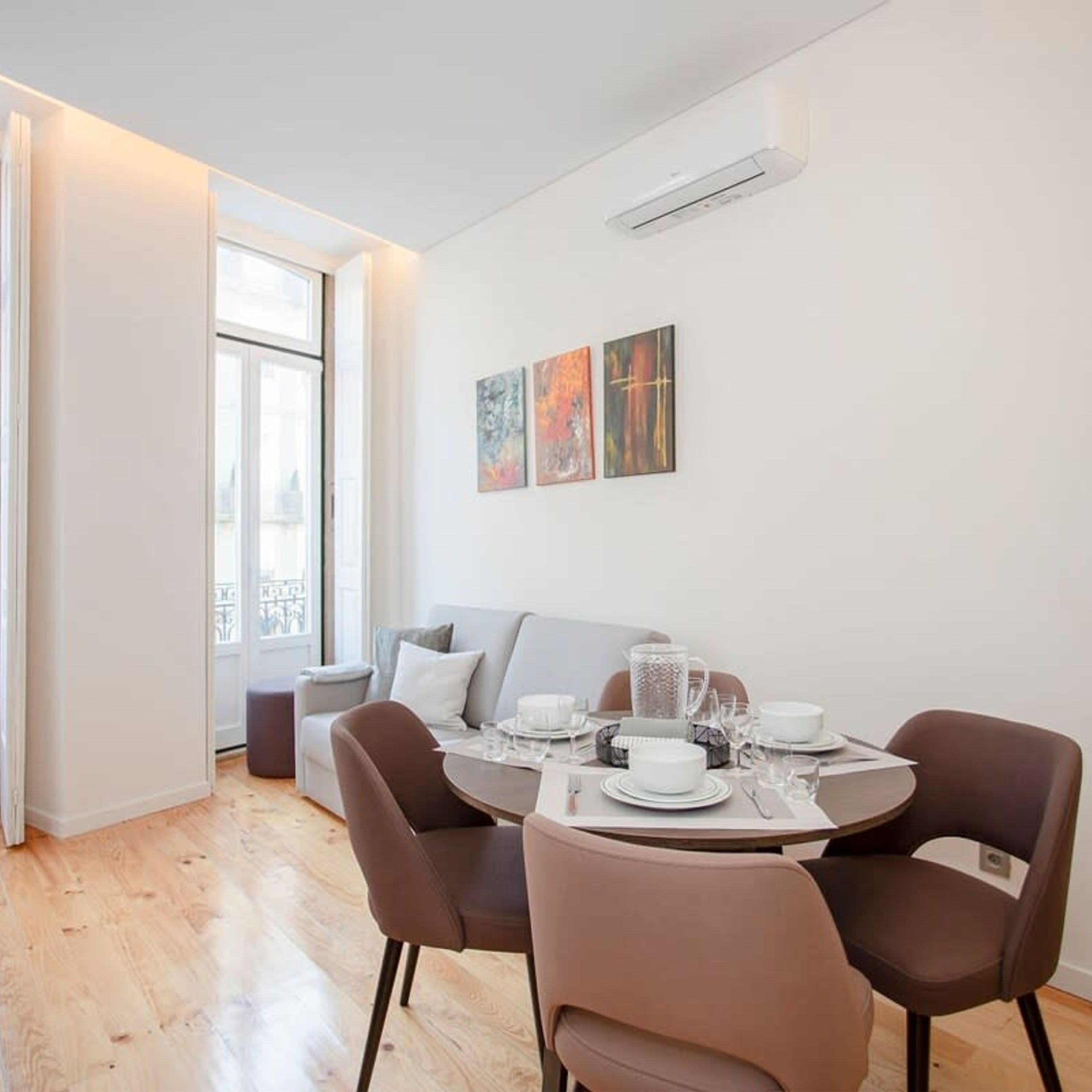 Un comedor moderno con una mesa redonda preparada para cuatro personas, un sofá gris y una ventana con vistas.