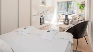 A modern hotel room with a large bed, a dining area with a table and chairs, a kitchen area, and a large window providing natural light. - Liiiving in Porto - City Design Apartment A (Porto)