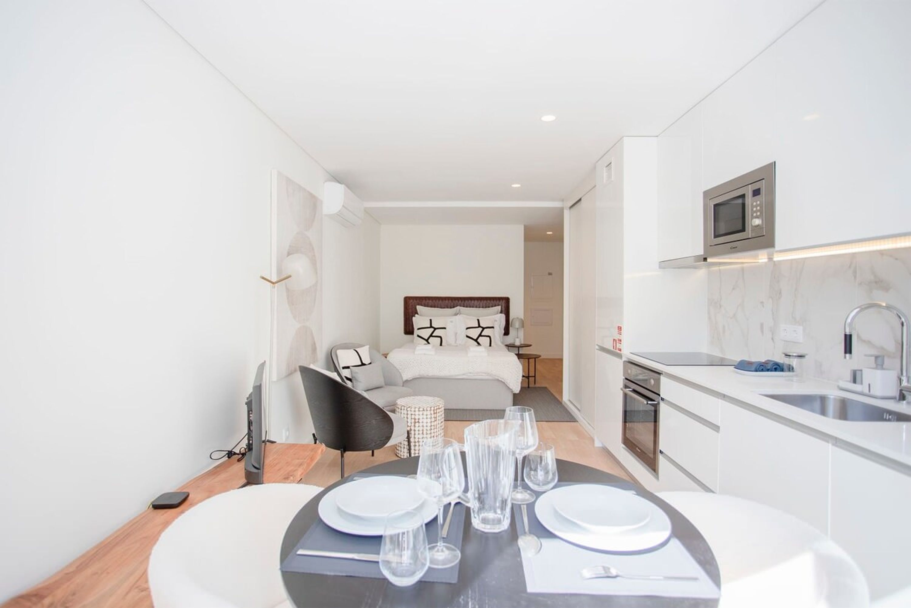 A modern, well-lit room with a dining table set for two, a bed with a headboard, a small kitchen area with a microwave, and a sink.