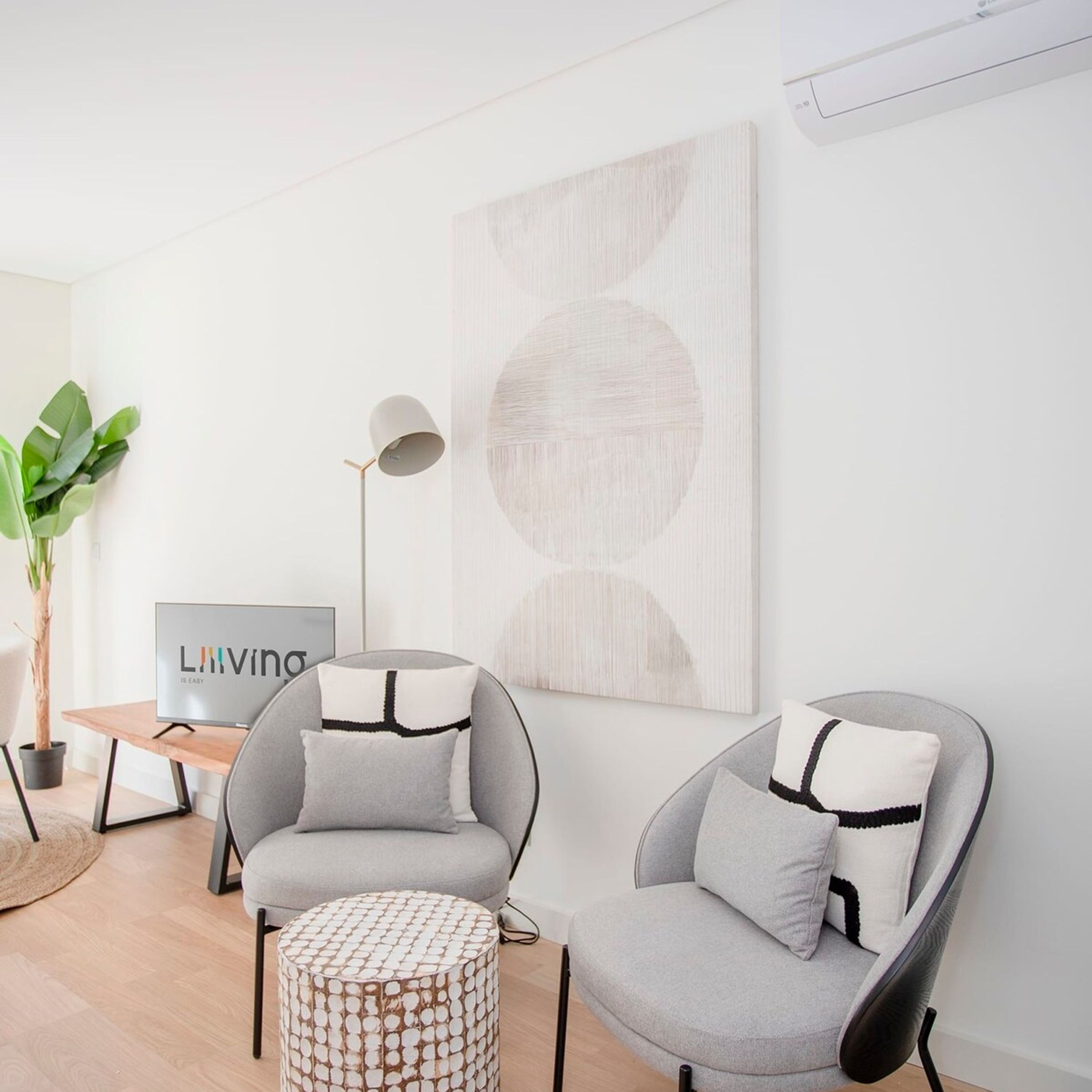 Un salon moderne avec deux fauteuils gris, un pouf rond, une lampe sur pied, un téléviseur et un grand tableau abstrait accroché au mur.