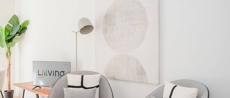 Un salon moderne avec deux fauteuils gris, un pouf rond, une lampe sur pied, un téléviseur et un grand tableau abstrait accroché au mur.
