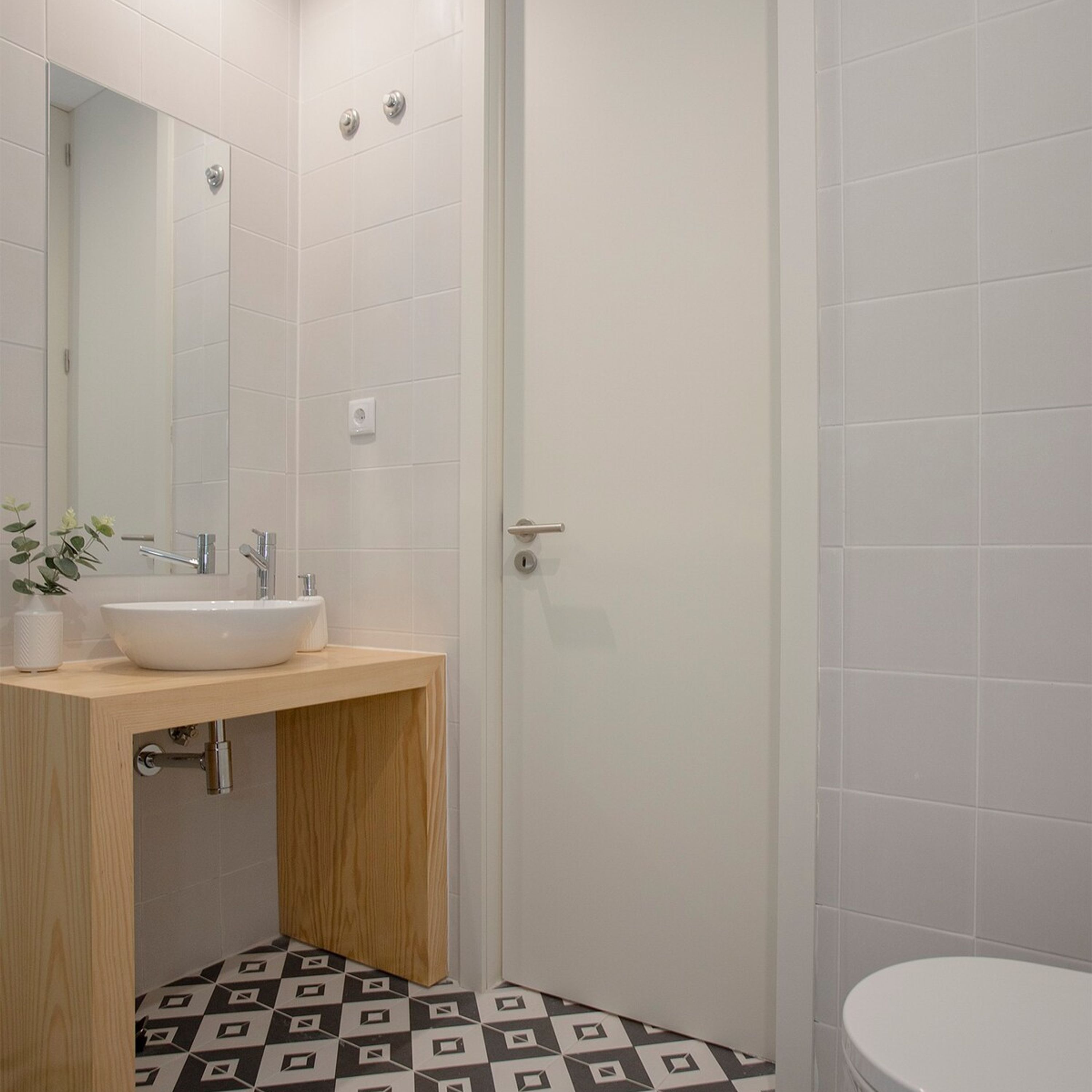 Et badeværelse med hvid vask, spejl og toilet. Gulvet har et sort-hvidt geometrisk mønster.
